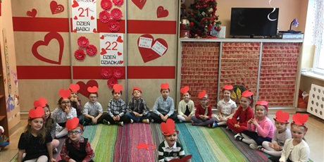 Powiększ grafikę: Dzieci  z  grupy O1 siedzą  na  podłodze. Na  głowach  maja  opaski  z  serduszkami. W  tle dekoracja  z  okazji Dnia Babci  i  Dziadka.