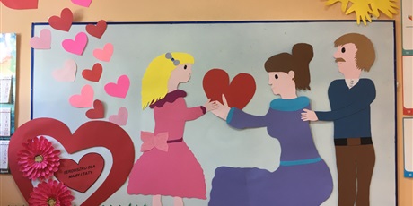 Powiększ grafikę: Na  tablicy  powieszona jest papierowa dekoracja, wycięta  rodzina, dziewczynka  podaje  serce  mamie.