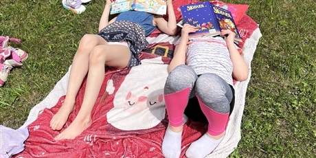 Powiększ grafikę: Na trawniku rozłożony jest koc, na którym, leżą uczennice, czytające książki.