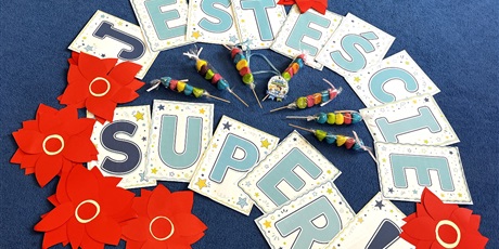 Powiększ grafikę: Na zdjęciu dekoracja z okazji Dnia Chłopaka. Na niebieskim tle leżą litery ułożone w napis "Jesteście super!". Po lewej stronie leżą czerwone kwiaty z papieru.