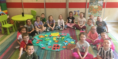 Powiększ grafikę: Grupa  dzieci  siedzi  w  kole  na  dywanie. Pośrodku  leży  ułożona  z  wyciętych  papierowych kwiatów  i  owadów, praca  plastyczna  z  napisem  "Majowa  łąka".