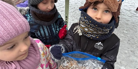 Powiększ grafikę: Przedszkolaki w zimowej scenerii z radością dokarmiają ptaki, trzymając ziarenka przygotowane do wsypania do karmnika w                             ogrodzie przedszkolnym.