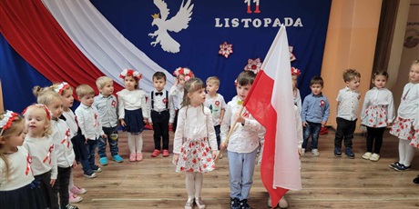 Powiększ grafikę: Grupa przedszkolaków  z flagą Polski stoi  na tle dekoracji z okazji 11 listopada.