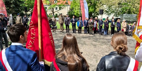 Powiększ grafikę: Po lewej stronie stoi uczeń z czerwonym sztandarem w dłoni. Po prawej obok niego stoją dwie uczennice z biało-czerwonymi szarfami przewieszonymi od prawego do lewego ramienia. Uczniowie patrzą na grupkę ludzi stojącą naprzeciwko nich, wśród której znajdują się  żołnierze w mundurach oraz mieszkańcy dzielnicy.