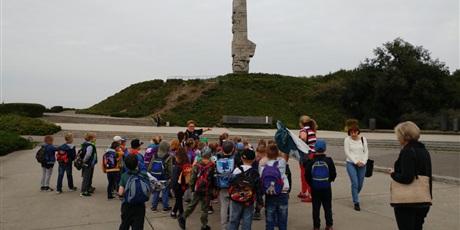 Powiększ grafikę: Grupa  dzieci  stoi tyłem  i  słucha  przewodnika. W  tle  widać  pomnik  na  Westerplatte.